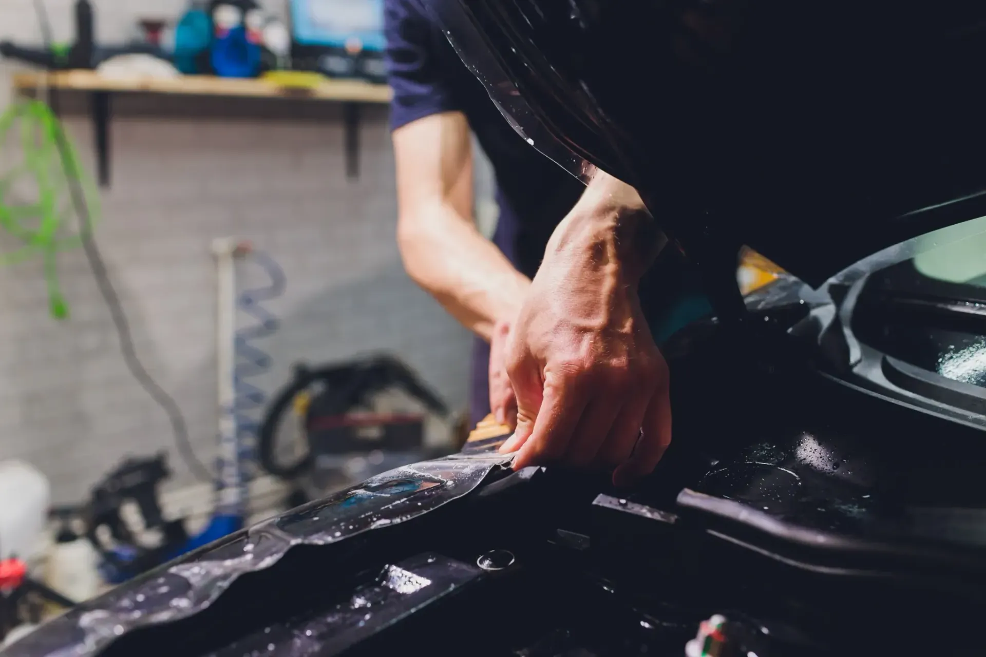 Persona aplicando película a un automóvil; manos cerca, trabajando en un vehículo oscuro en un taller.