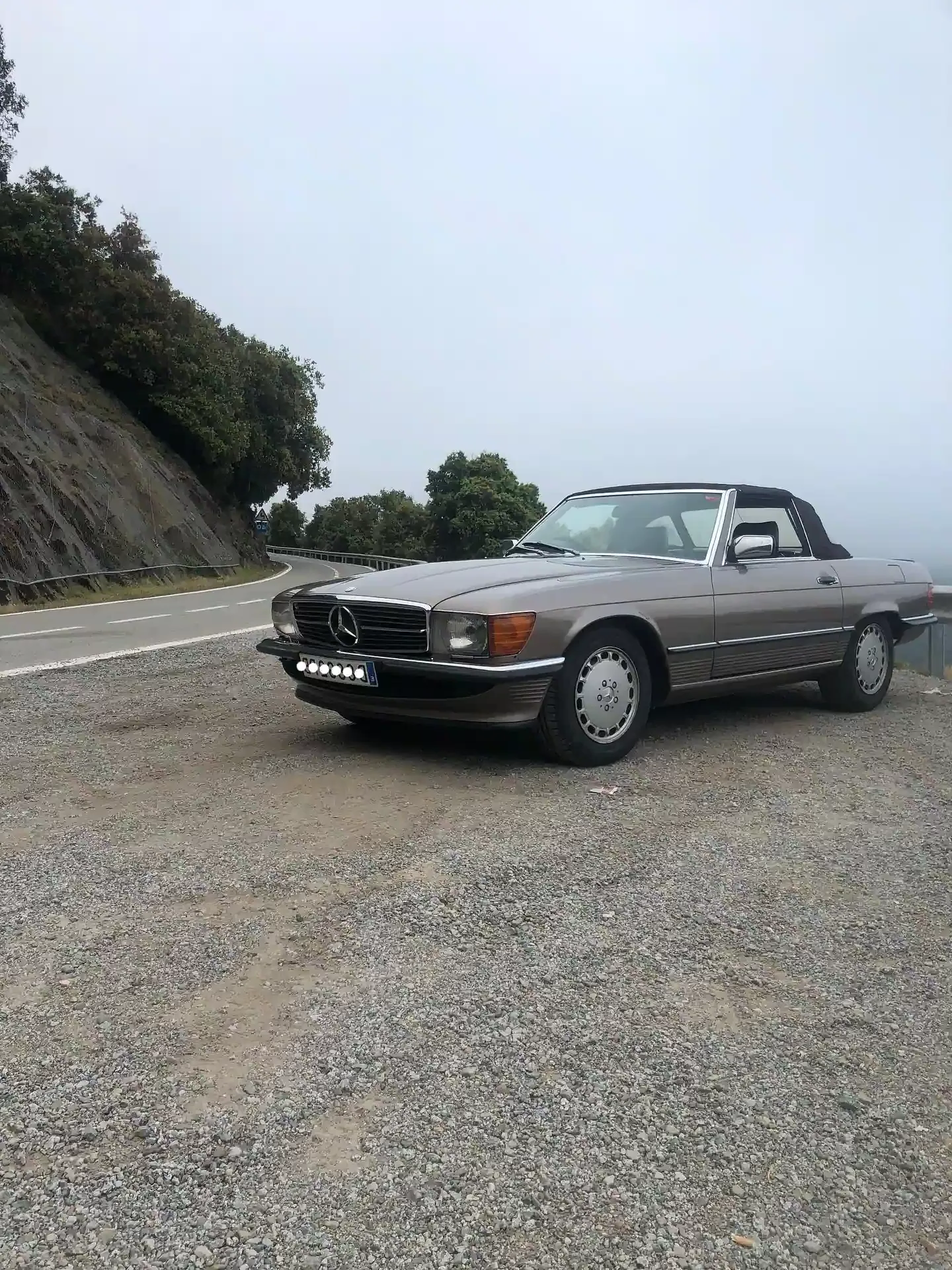 Mercedes-Benz convertible de color beige estacionado en un arcén de grava cerca de una carretera, día nublado.
