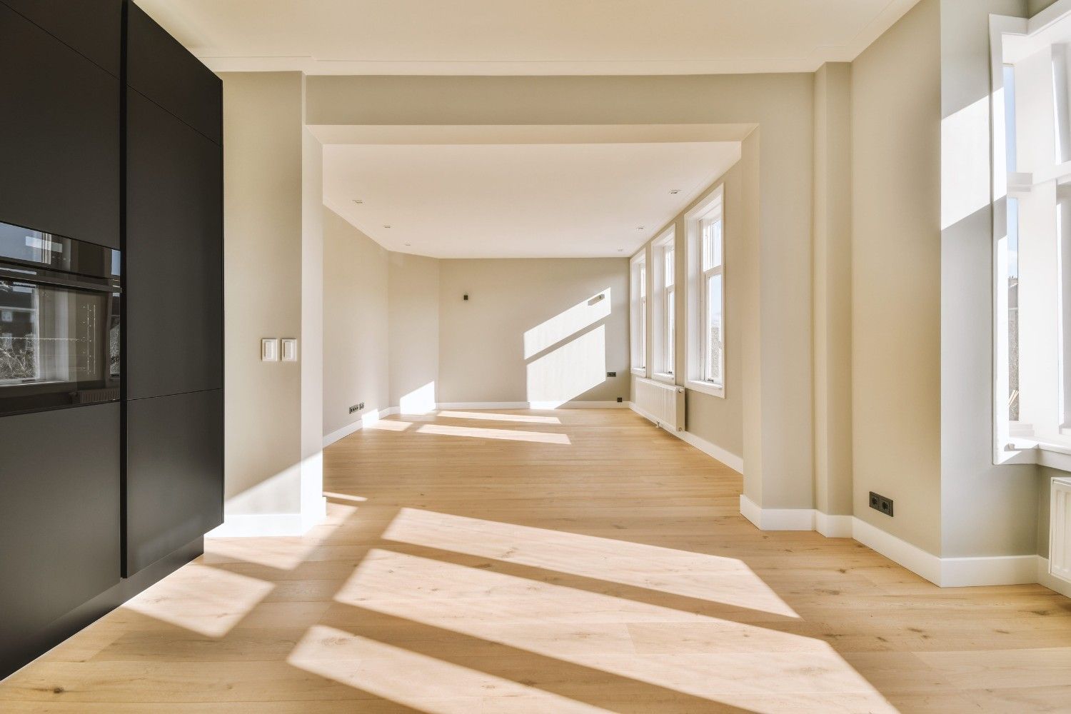 Sala de estar luminosa y vacía con suelos de madera clara y luz solar que entra a raudales por los grandes ventanales.