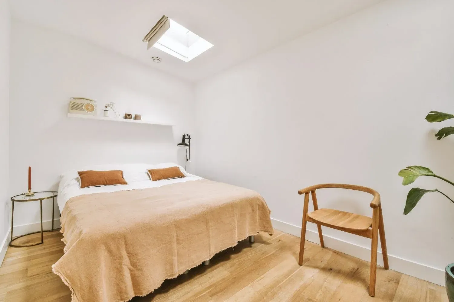 Dormitorio luminoso y minimalista con cama beige, suelo de madera, tragaluz y silla de madera cerca de una planta frondosa.