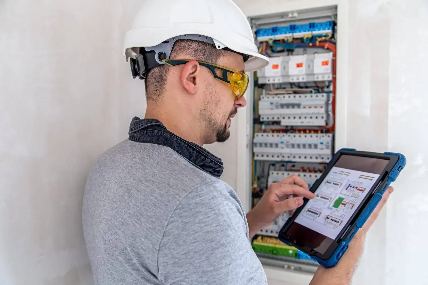 Técnico con casco y gafas de seguridad utilizando una tableta para inspeccionar un panel eléctrico.