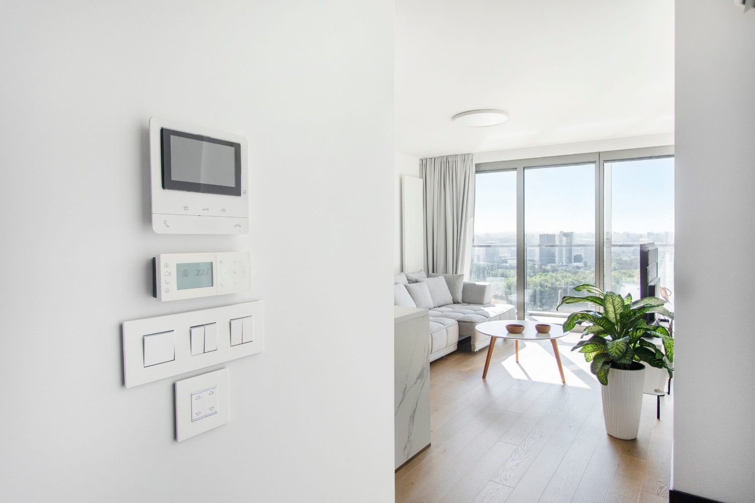 Luminoso salón con controles en la pared, sofá, sillón y planta en maceta junto a una gran ventana con vistas a la ciudad.