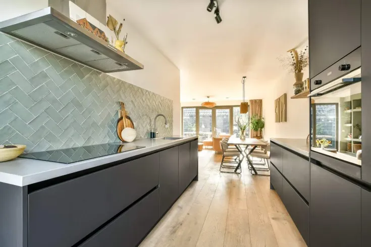 Cocina moderna con gabinetes oscuros, salpicadero gris y zona de comedor contigua, iluminada por luz natural.