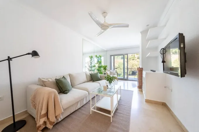Luminoso salón con sofá blanco, mesa de centro de cristal, televisor montado en la pared y puertas que dan al balcón.