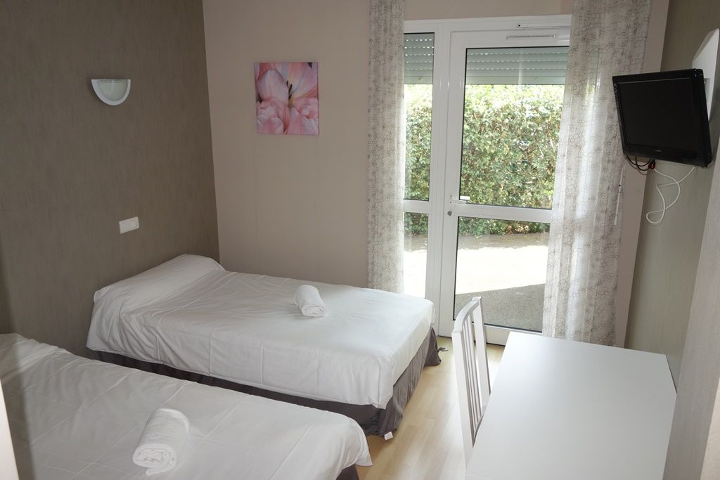 Petite chambre avec deux lits, un bureau blanc, des œuvres d’art murales et des portes-fenêtres avec des rideaux