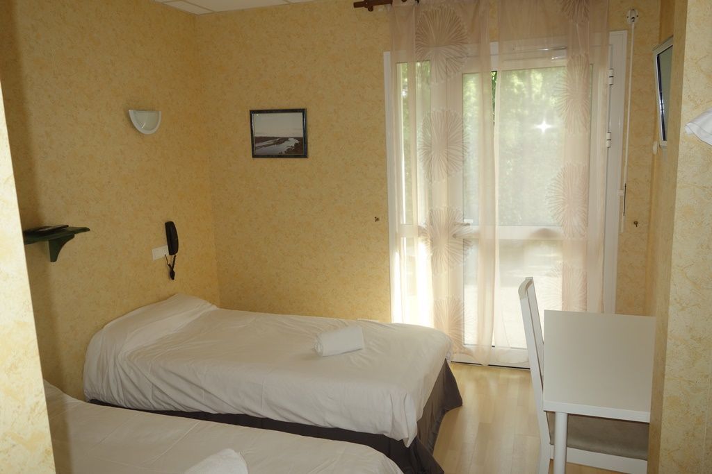 Petite chambre avec un lit simple, un bureau et une fenêtre lumineuse ornée de voilages.
