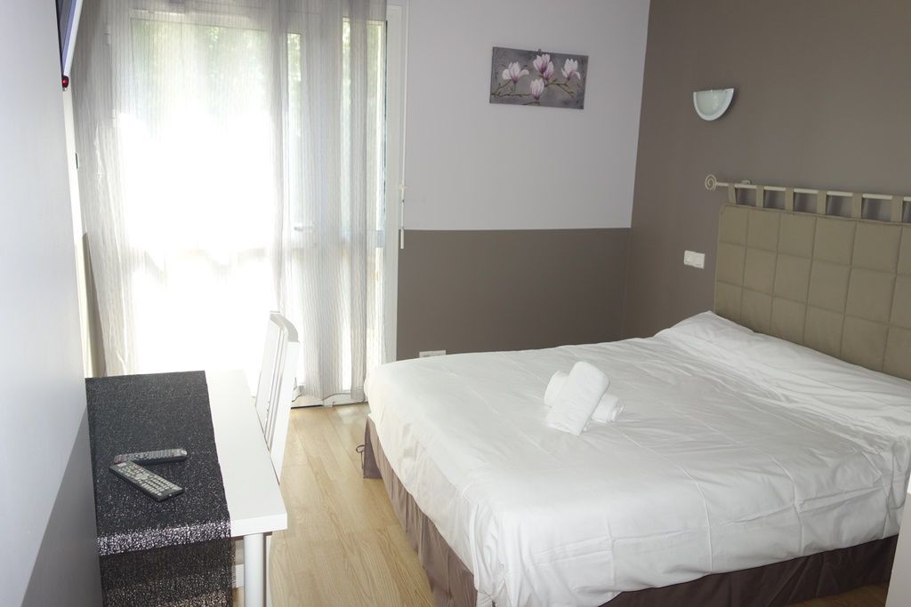 Chambre d’hôtel lumineuse avec un lit double, un bureau, une chaise et une grande fenêtre avec des rideaux