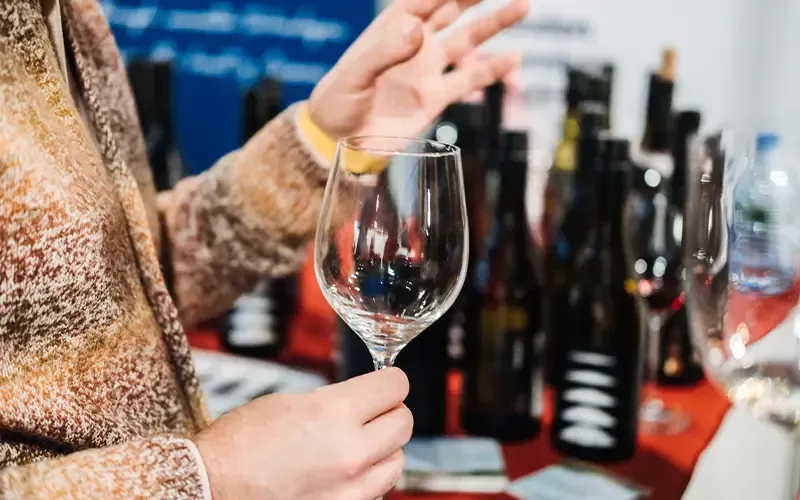 Personne tenant un verre de vin devant un présentoir de dégustation, avec des bouteilles en arrière-plan