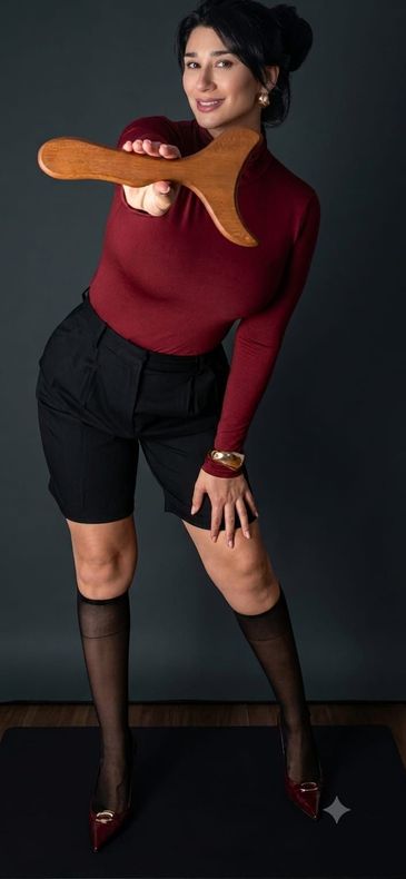 Mujer posando con top burdeos, pantalones cortos negros y medias, sosteniendo un objeto con forma de pretzel sobre un fondo oscuro.
