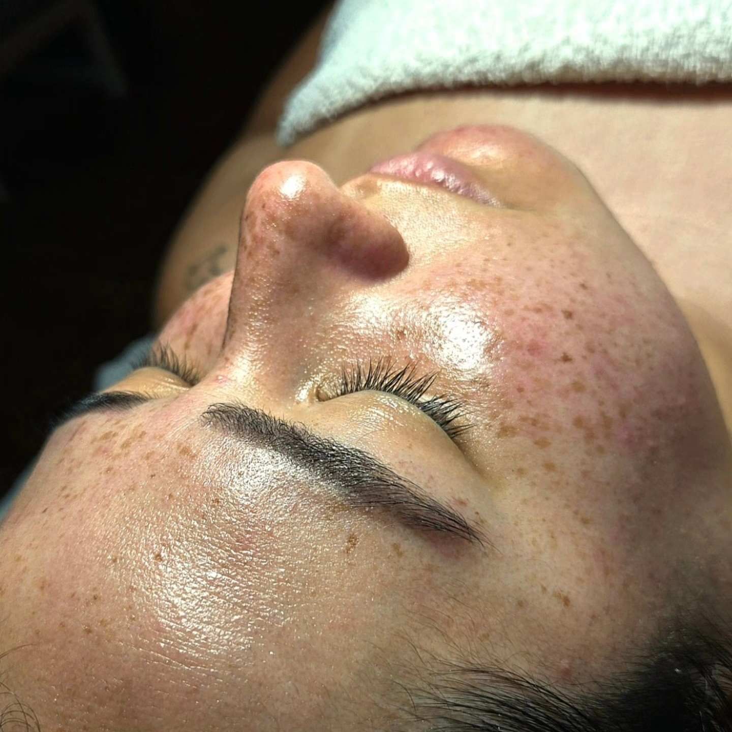 Primer plano del rostro de una persona con un tratamiento facial brillante, con los ojos cerrados, tumbada en una camilla de spa.