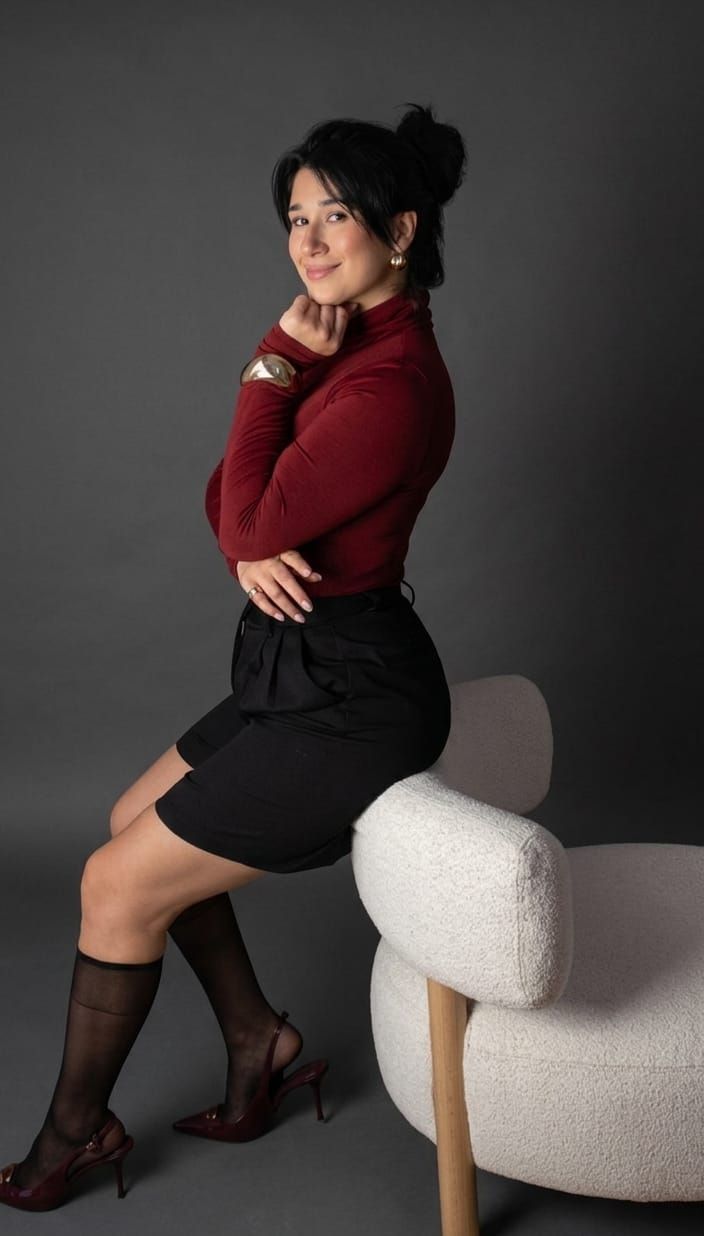 Mujer sentada en una silla moderna, con una blusa roja y una falda negra, frente a un fondo de estudio oscuro.