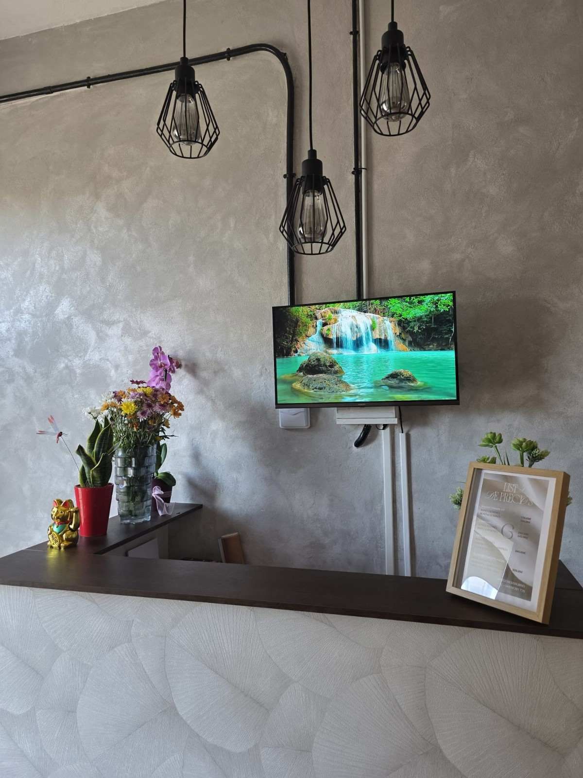 Mostrador de recepción con flores, cartel enmarcado, luces colgantes y un televisor que muestra una cascada en un vestíbulo moderno.