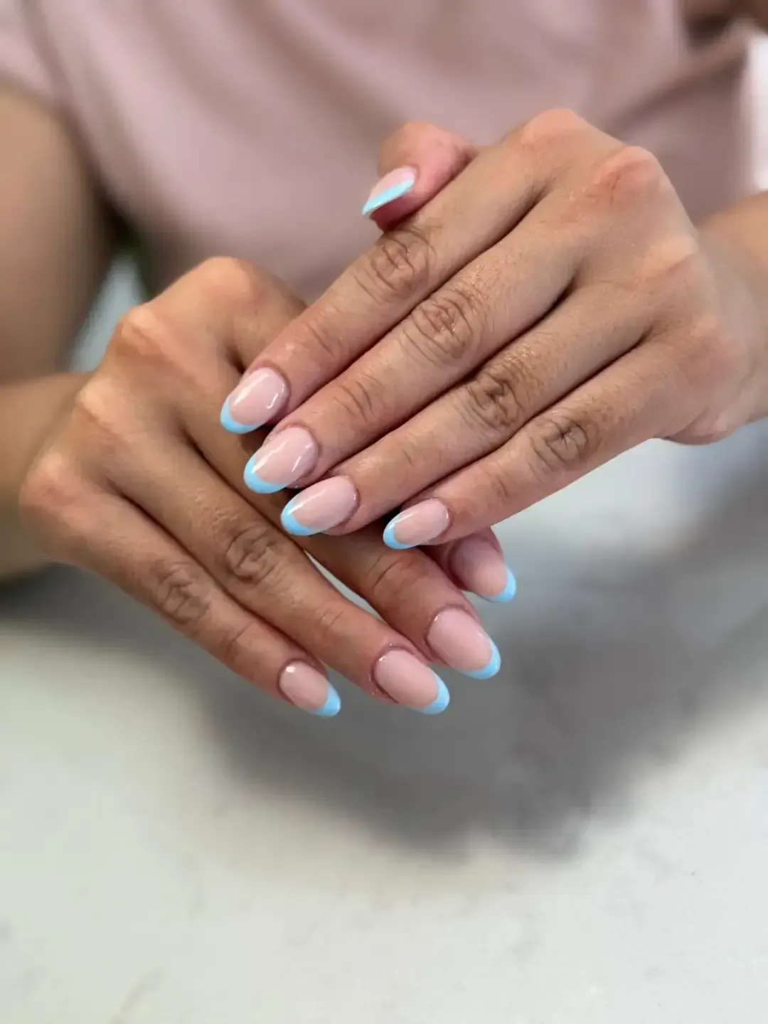 Manos con uñas ovaladas con base rosa claro y puntas francesas azules, descansando sobre una superficie blanca.