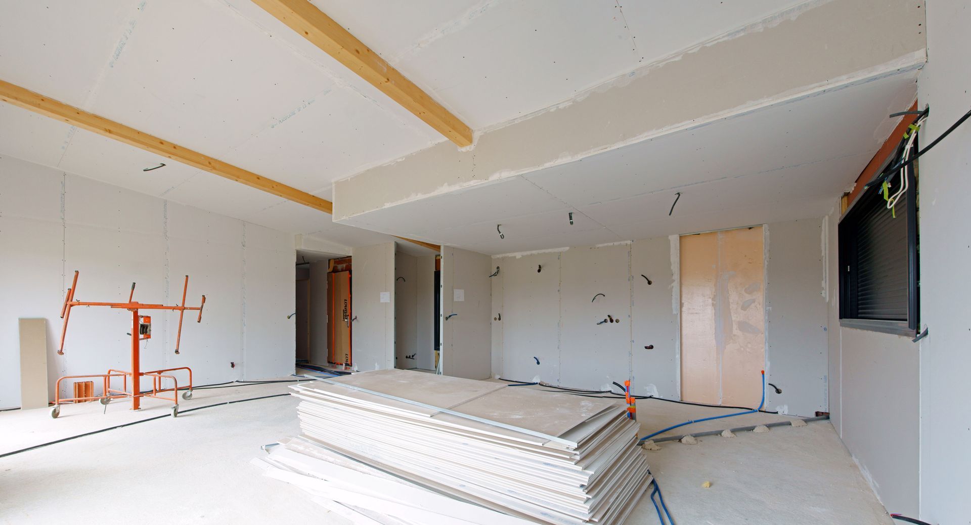 Pièce inachevée avec plaques de plâtre empilées, charpente apparente et murs blancs en cours de construction.