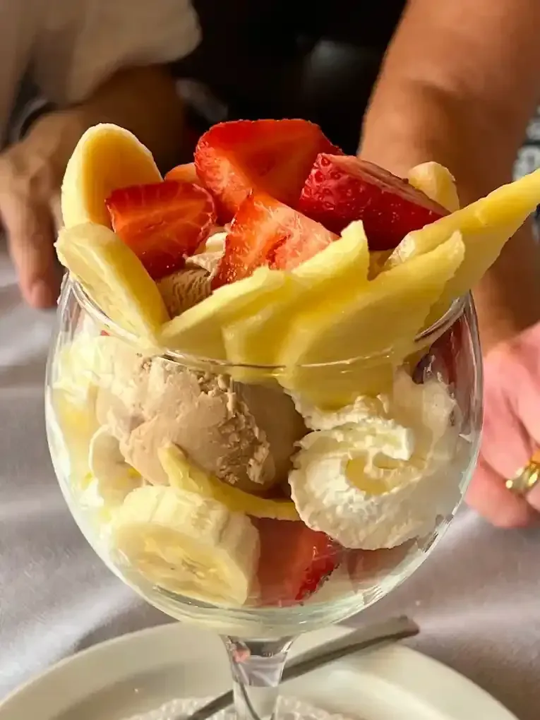 Vaso de postre de cristal con rodajas de plátano, fresa y mango dispuestas en capas sobre bolas de helado.