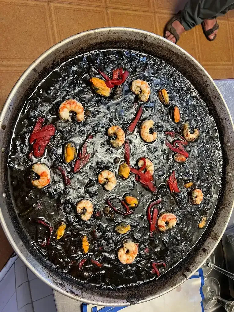 Sartén de arroz negro o estofado cubierto con camarones, pimientos rojos y hierbas sobre un piso de baldosas.