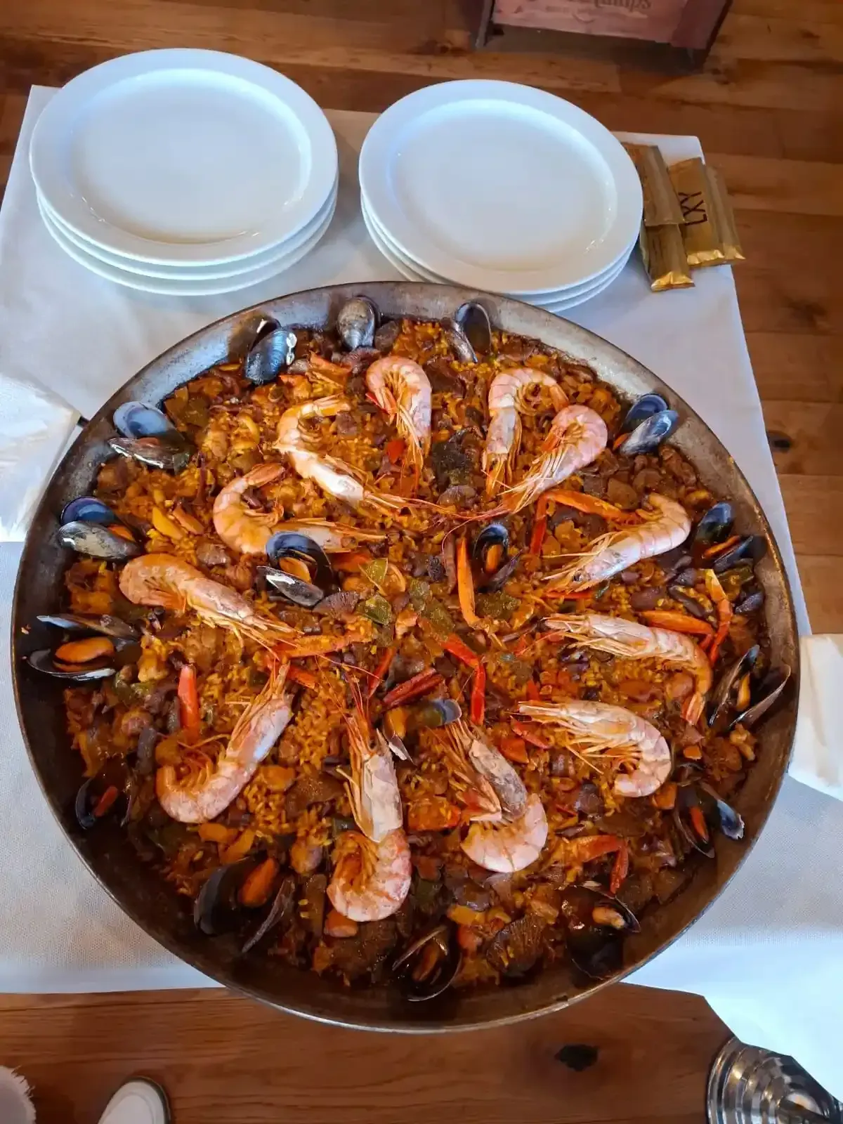 Una paellera con gambas y mejillones sobre una mesa de madera, con dos cuencos blancos cerca.
