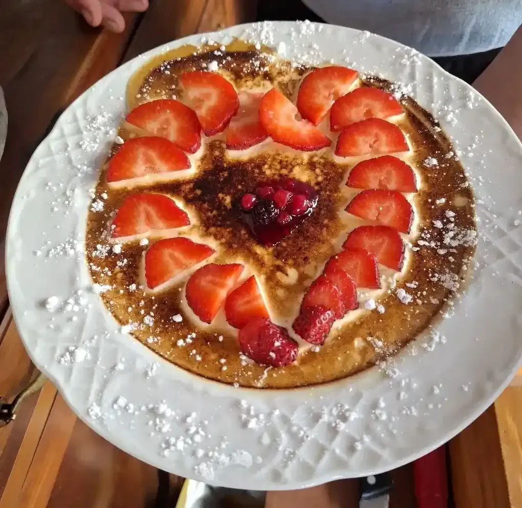 Tortita con forma de corazón cubierta con fresas en rodajas, azúcar glas y salsa de frutos rojos sobre un plato blanco.