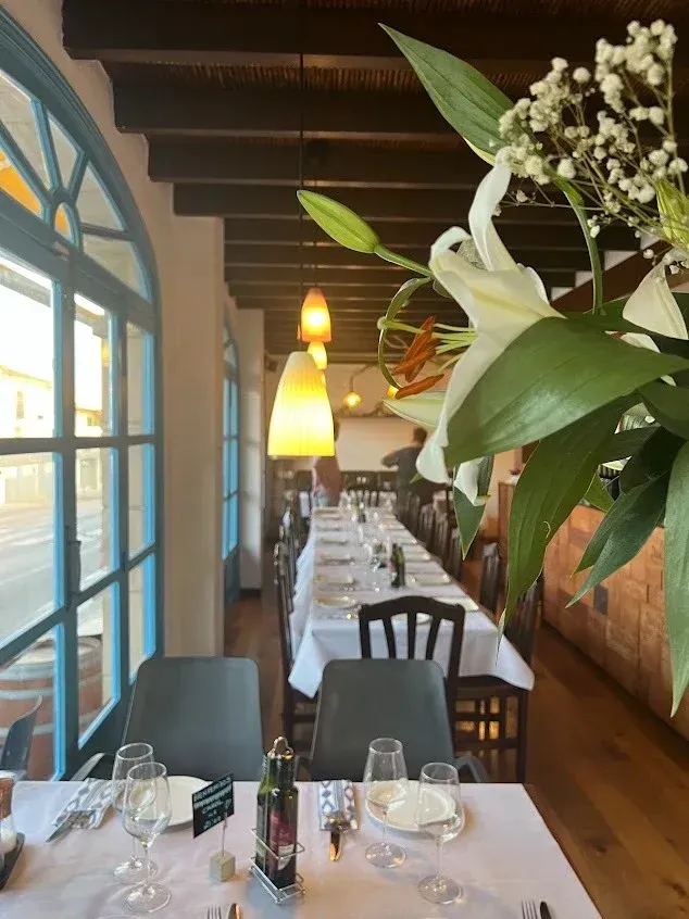 Acogedor comedor de restaurante con mesas puestas, ventanas azules, cálidas lámparas colgantes y centro de mesa floral.