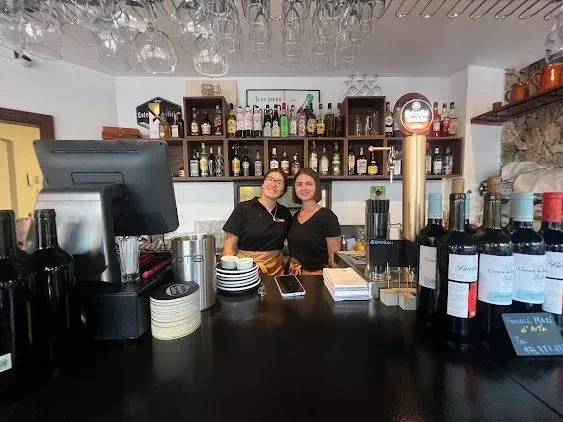 Dos personas detrás de la barra de un bar con botellas y vasos expuestos en estantes detrás de ellas.