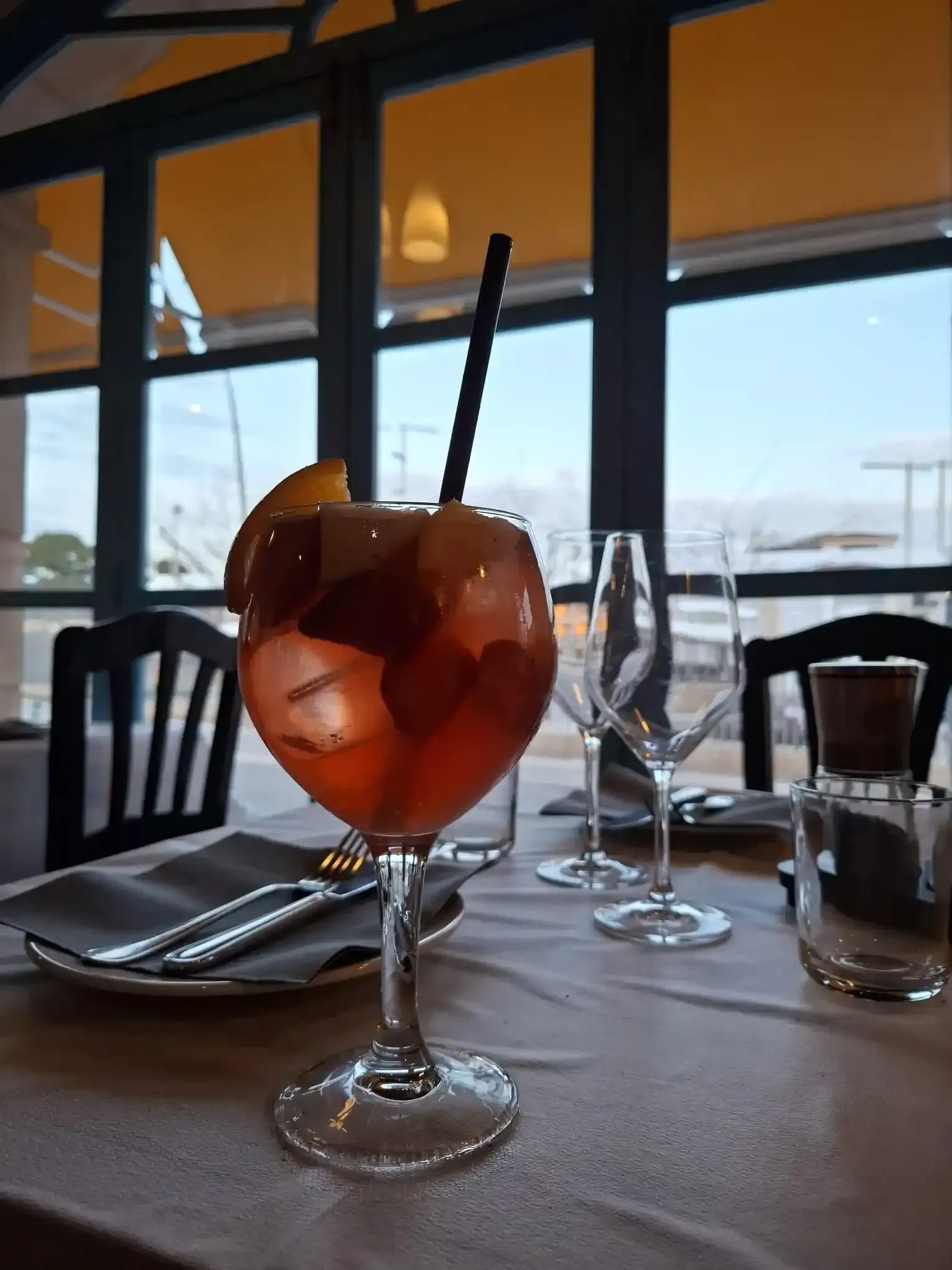 Un cóctel servido en una copa con tallo y rodajas de naranja sobre una mesa junto a una ventana con vistas al agua.