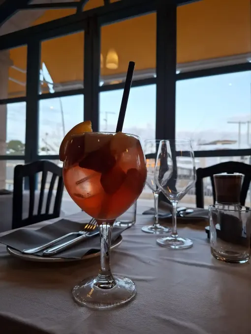Un cóctel servido en una copa con tallo y rodajas de naranja sobre una mesa junto a una ventana con vistas al agua.