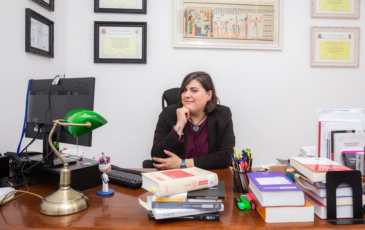 Una mujer con un blazer negro está sentada en un escritorio en una oficina. Sonríe, apoyando la barbilla en la mano. Se ven libros, una computadora y una lámpara de escritorio verde.