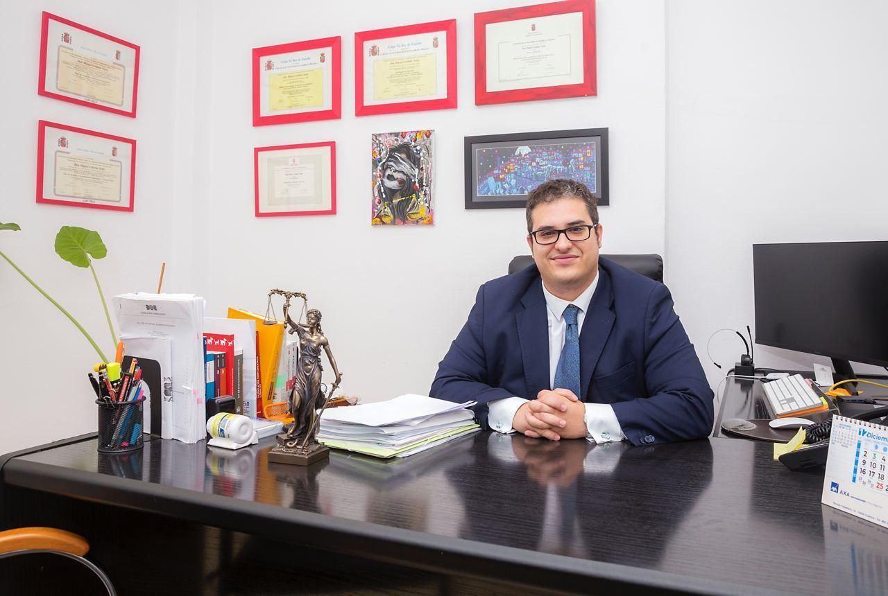 Un hombre de traje está sentado en un escritorio en una oficina, mirando a la cámara. Detrás de él, hay diplomas y obras de arte colgados en la pared.