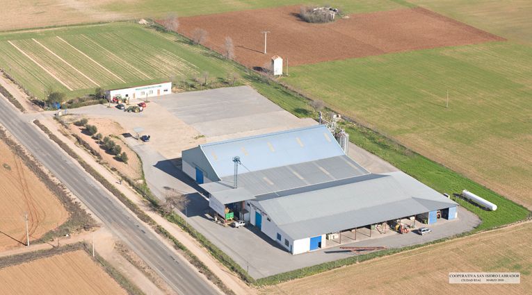 Vista aérea de un edificio agrícola con techo gris junto a caminos de tierra y campos.