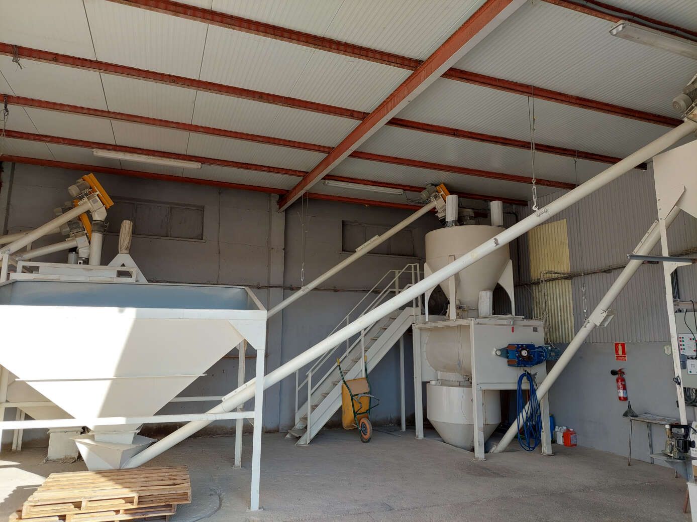 Nave industrial con maquinaria de transporte y escalera bajo un techo de chapa ondulada.