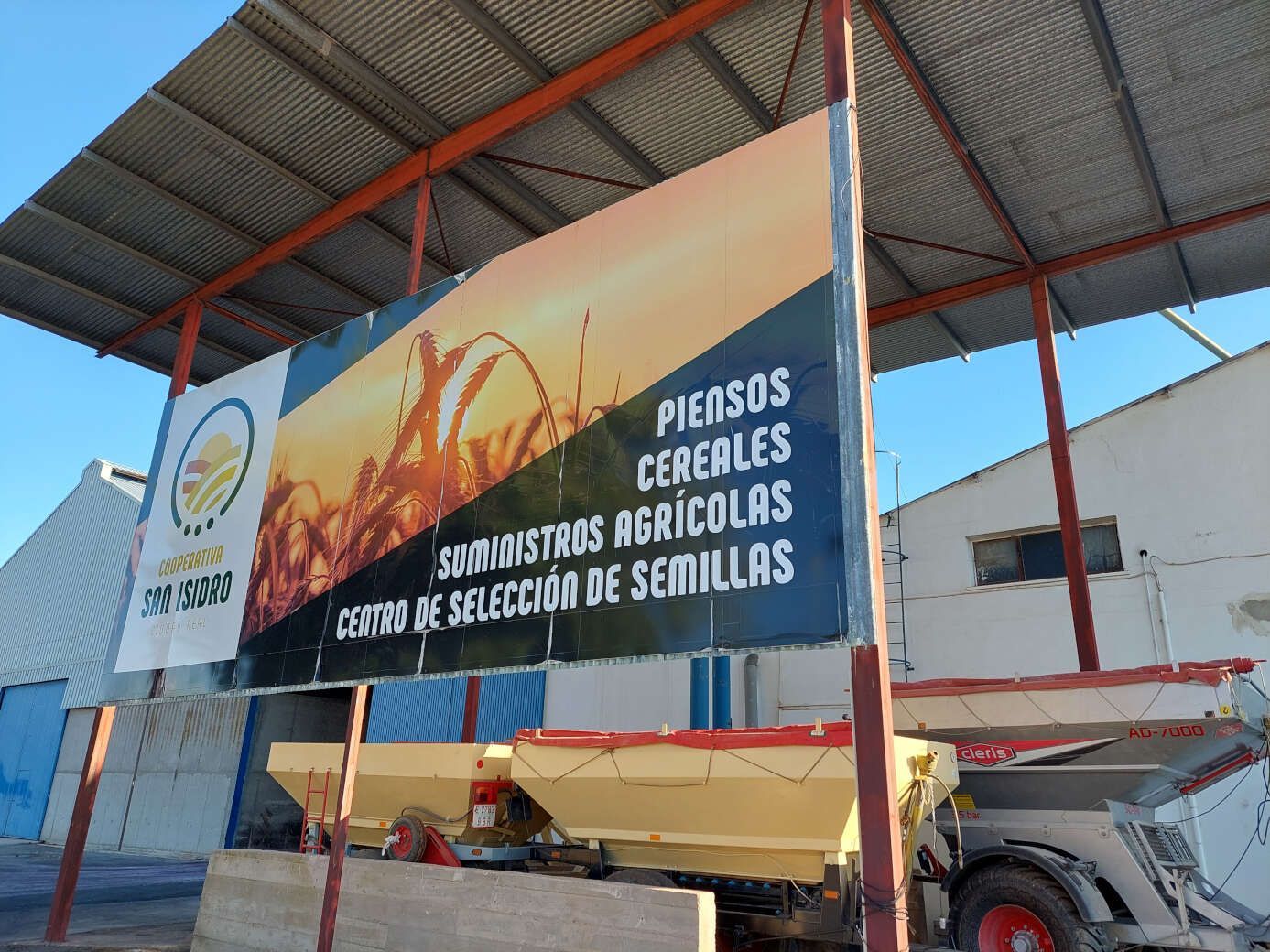 Gran pancarta que anuncia una tienda de semillas agrícolas bajo techo, con texto en español y maquinaria agrícola en las cercanías.