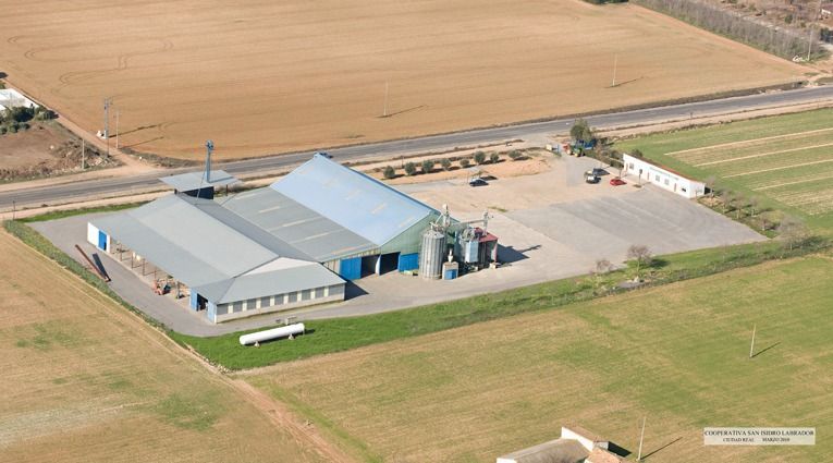 Vista aérea de un edificio con techo gris junto a campos y una zona de aparcamiento pavimentada en un entorno rural.