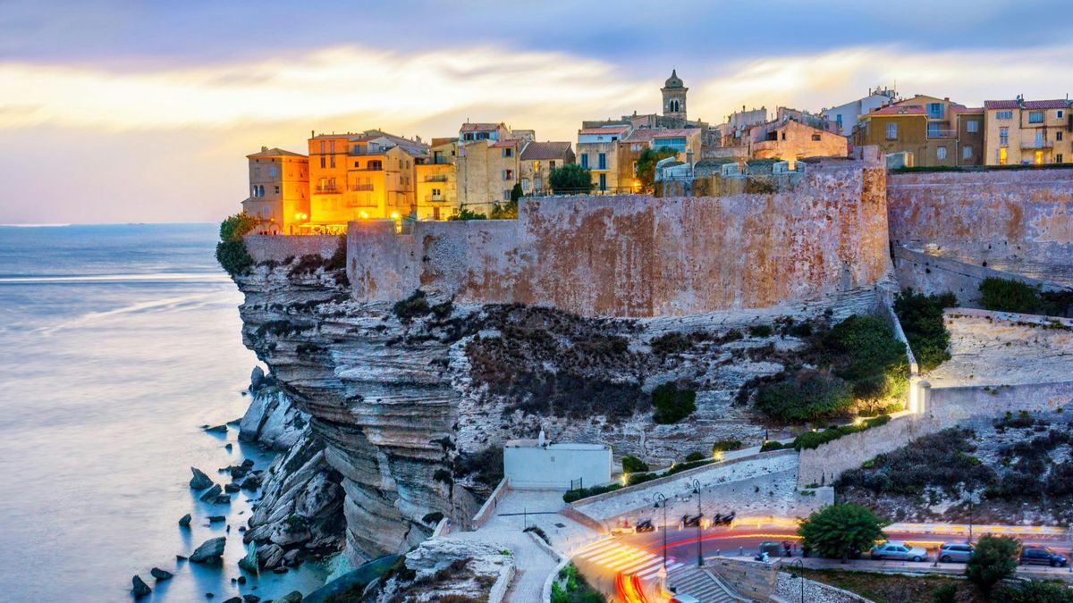 Ciudad acantilada iluminada al anochecer, con vistas al mar. Se ven edificios, murallas y una carretera costera.