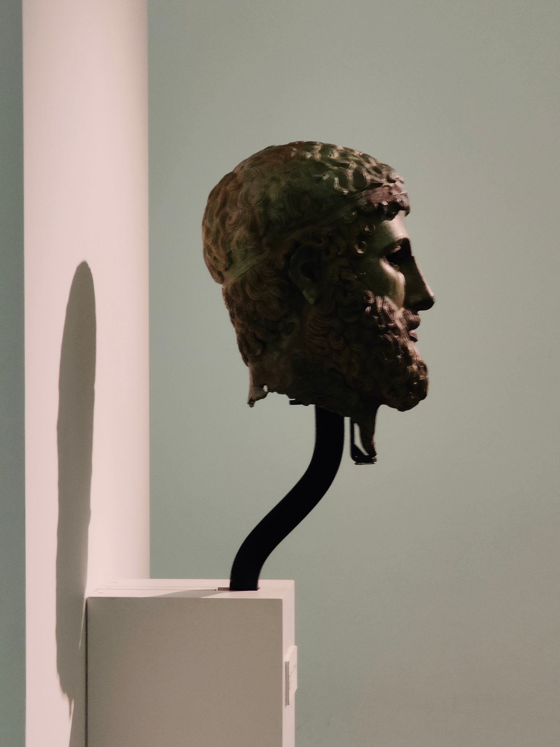 Escultura de cabeza de bronce de un hombre barbudo de perfil sobre un soporte negro, expuesta contra una pared blanca.