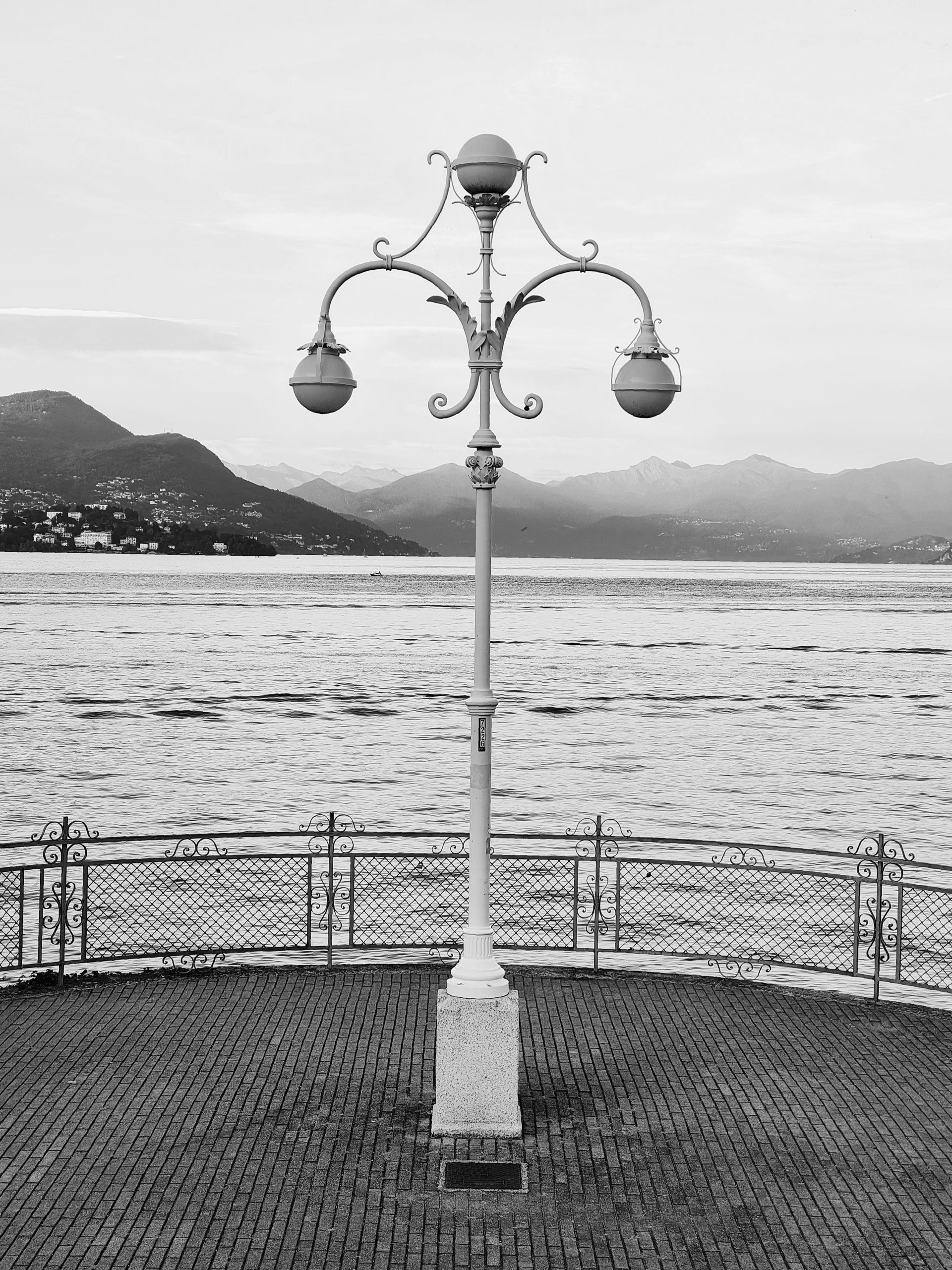 Fotografía en blanco y negro de una farola ornamentada en un paseo marítimo, con montañas y agua al fondo.