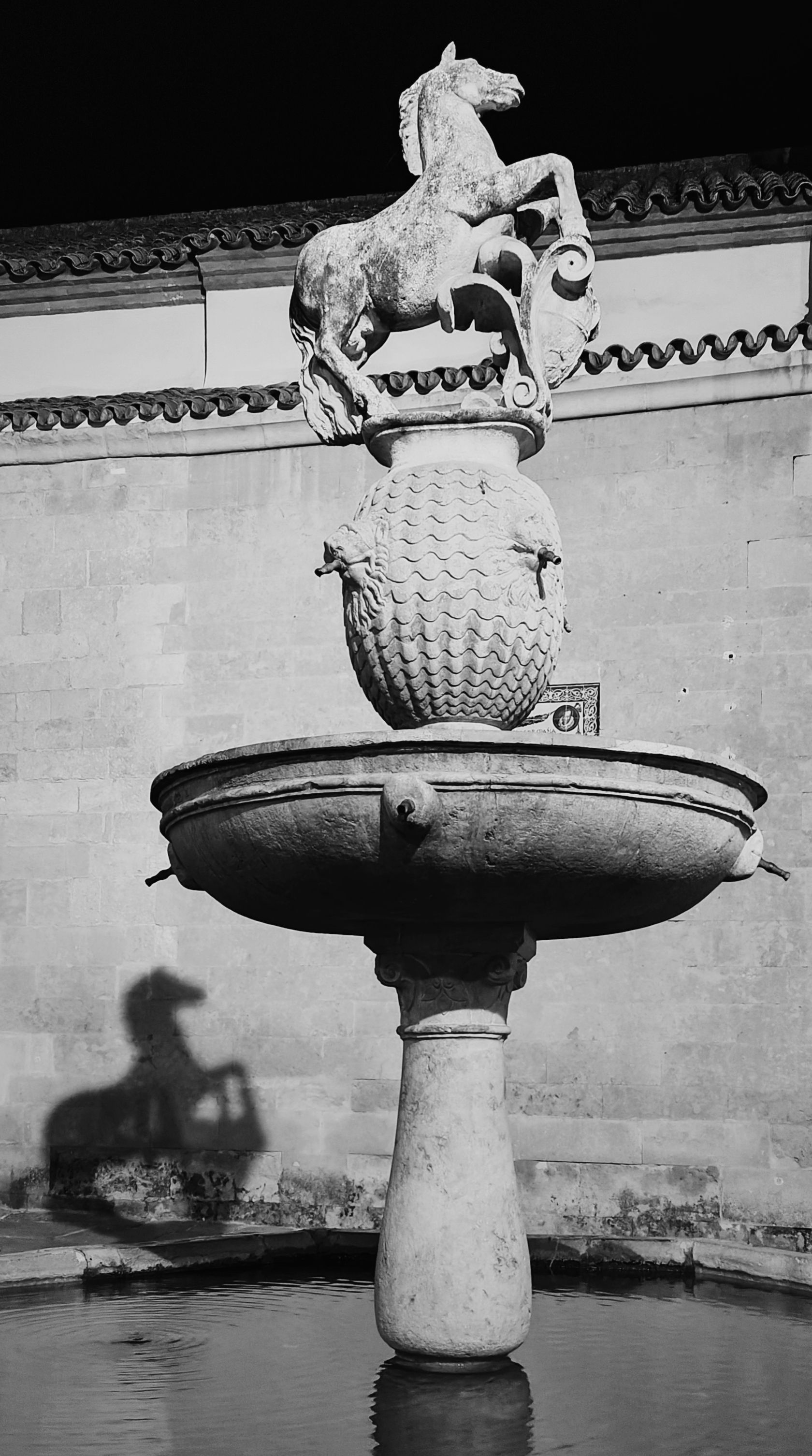 Fuente de piedra con estatua de caballo en la parte superior, proyectando una sombra.