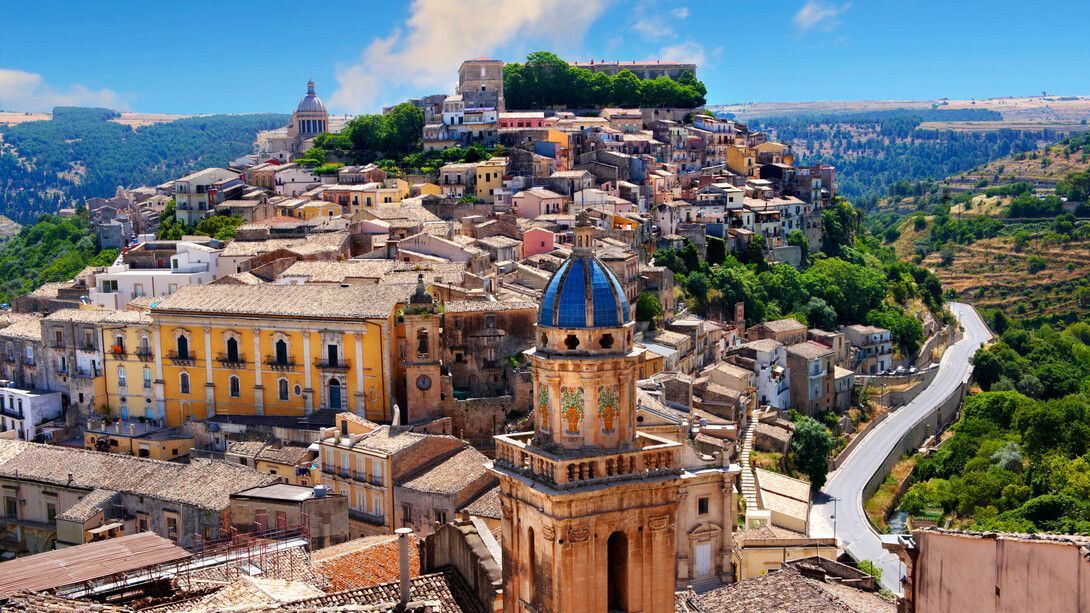 Ciudad italiana en la cima de una colina con iglesias, edificios coloridos y carreteras sinuosas bajo un cielo azul.