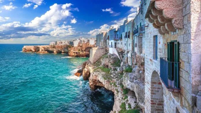 Pueblo costero con edificios blancos y azules sobre un acantilado con vistas al mar turquesa bajo un cielo azul.