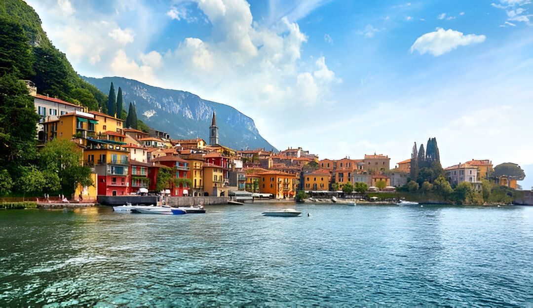 Edificios coloridos junto a un lago, bajo un cielo azul brillante. Barcos en el agua, montaña al fondo.