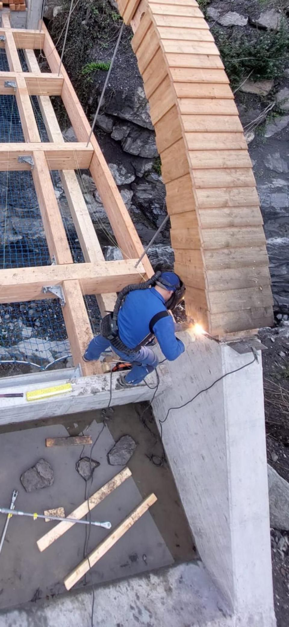 Un homme soude une structure en bois sur un pilier en béton.
