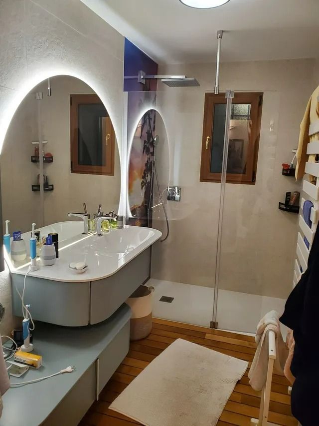 Petite salle de bains avec miroir ovale, lavabo, douche et carrelage beige au sol.