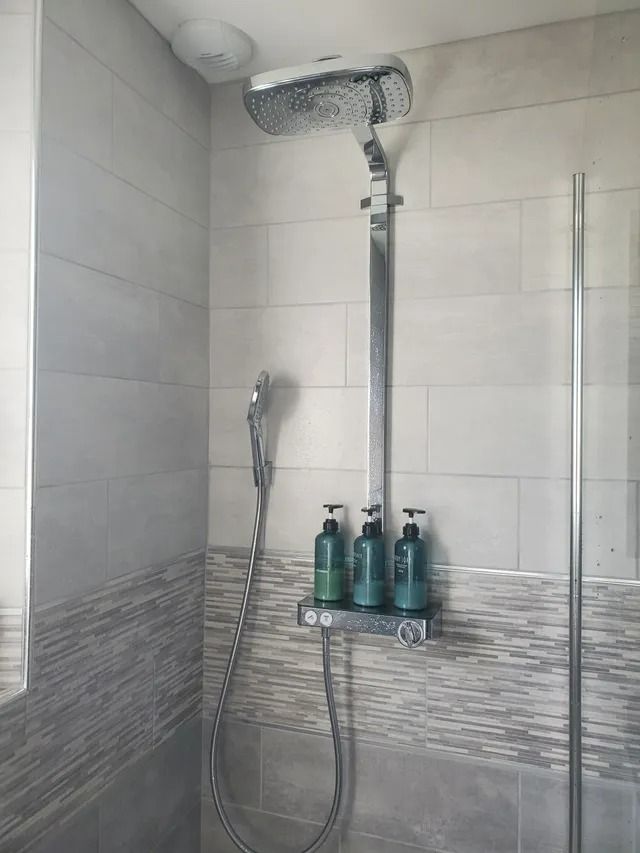 Douche à l'italienne avec pommeau effet pluie, douchette et trois flacons de shampoing verts sur une étagère.