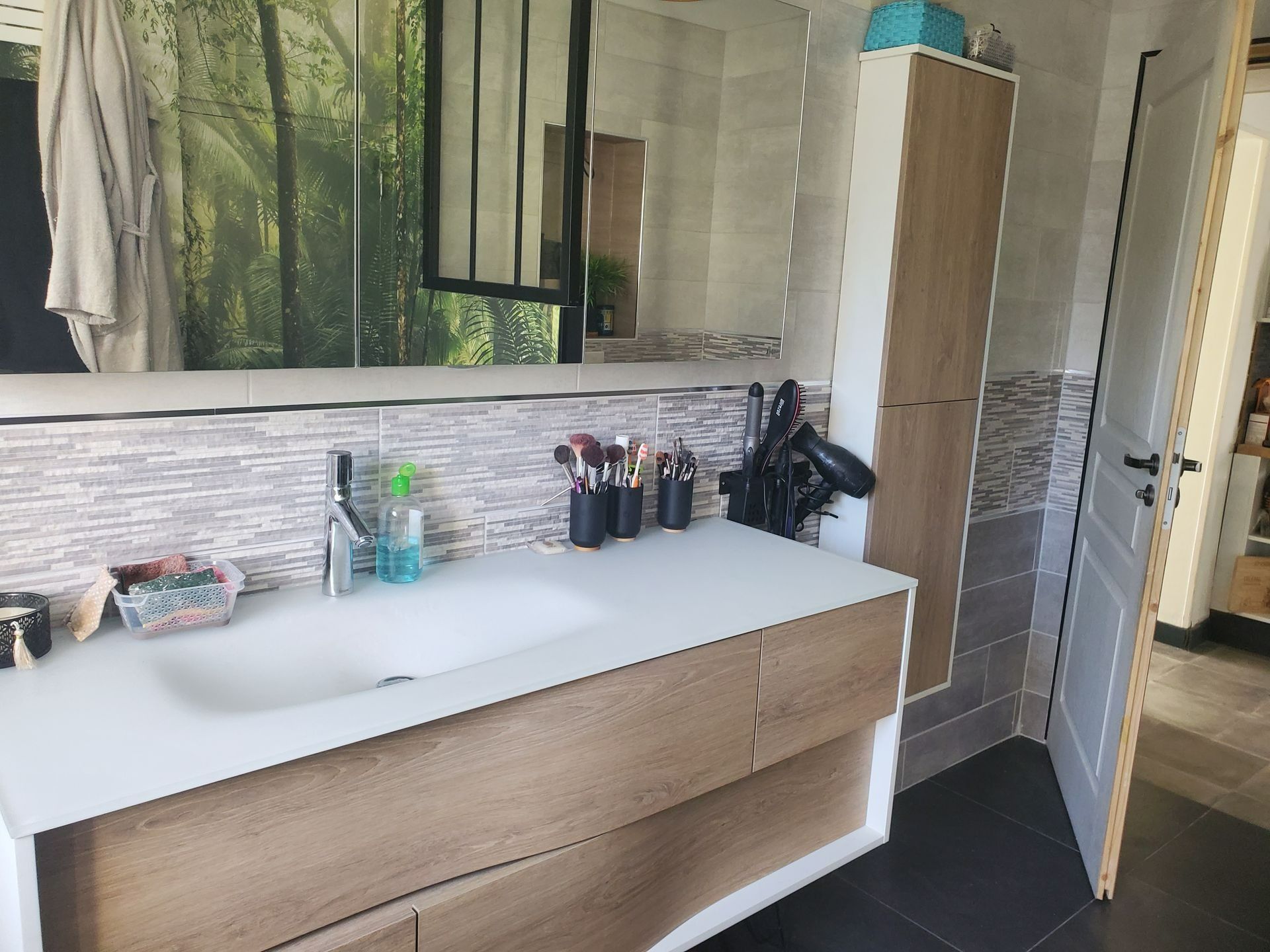 Meuble-lavabo moderne avec lavabo blanc, armoires en bois, miroir, articles de toilette et porte ouverte sur un espace douche.
