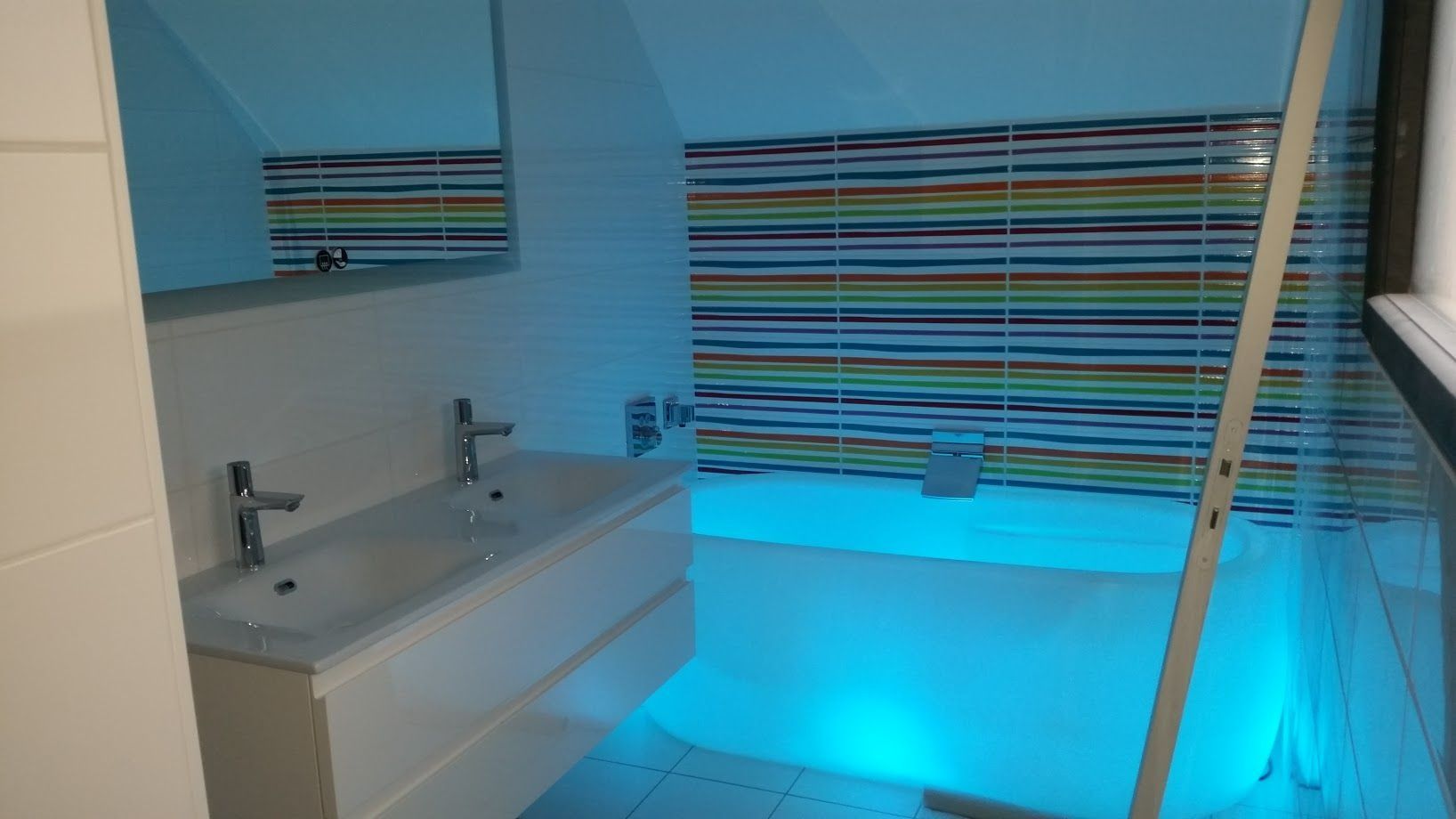 Salle de bains moderne avec lavabo blanc, stores rayés et éclairage bleu au-dessus de la baignoire.