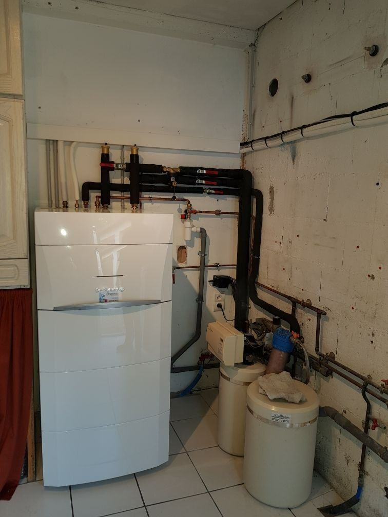 Buanderie avec une chaudière blanche, des tuyaux et un grand appareil électroménager blanc contre un mur éraflé.