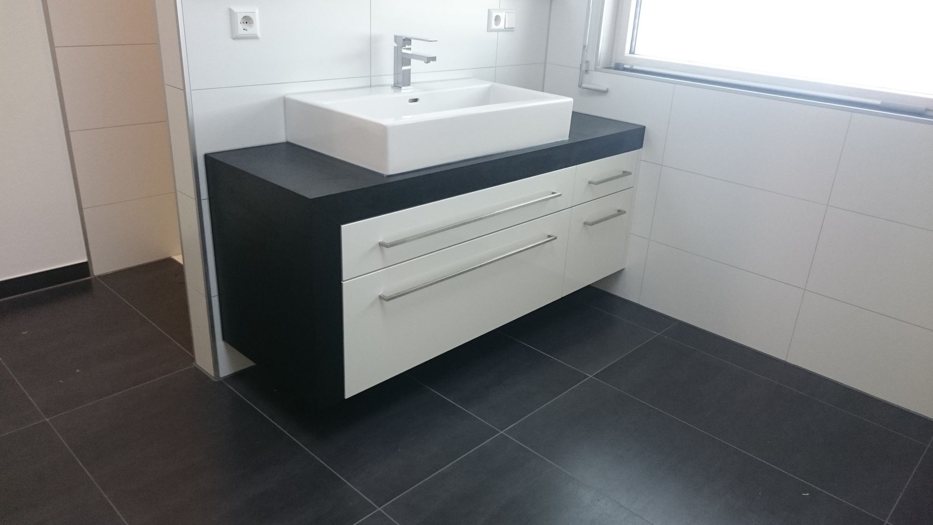 Modernes weißes Badezimmerwaschbecken und schwarze Arbeitsplatte vor weiß gefliesten Wänden und dunklem Boden.