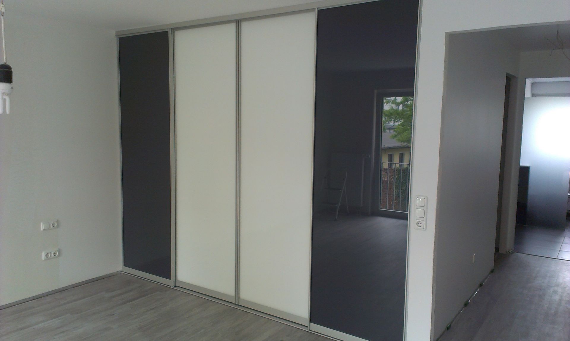 Leeres, modernes Zimmer mit weißen Wänden, grauem Boden und großen, dunklen Schiebetüren zum Kleiderschrank.