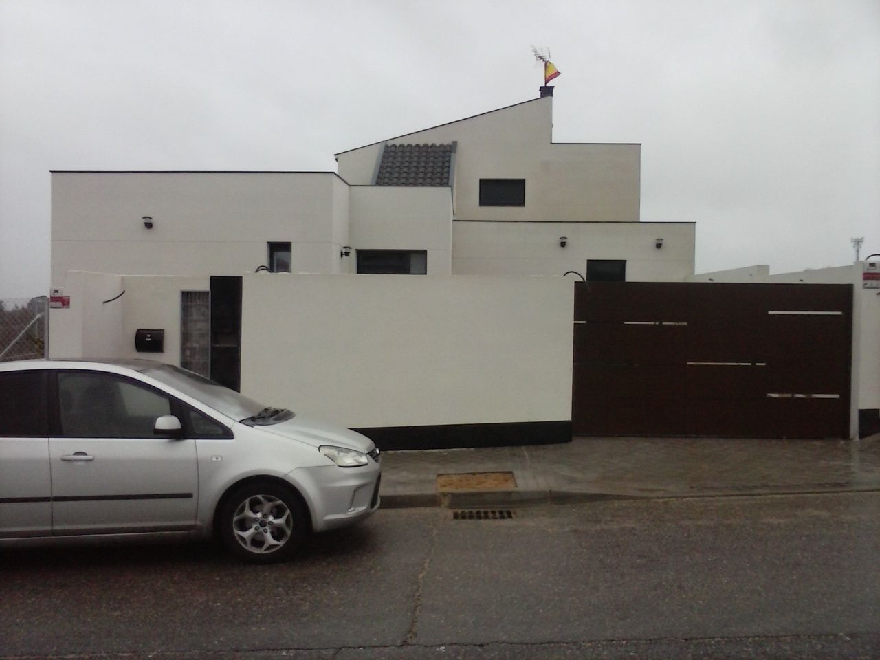Casa blanca moderna con portón marrón oscuro y coche plateado aparcado delante. Cielo gris y nublado.