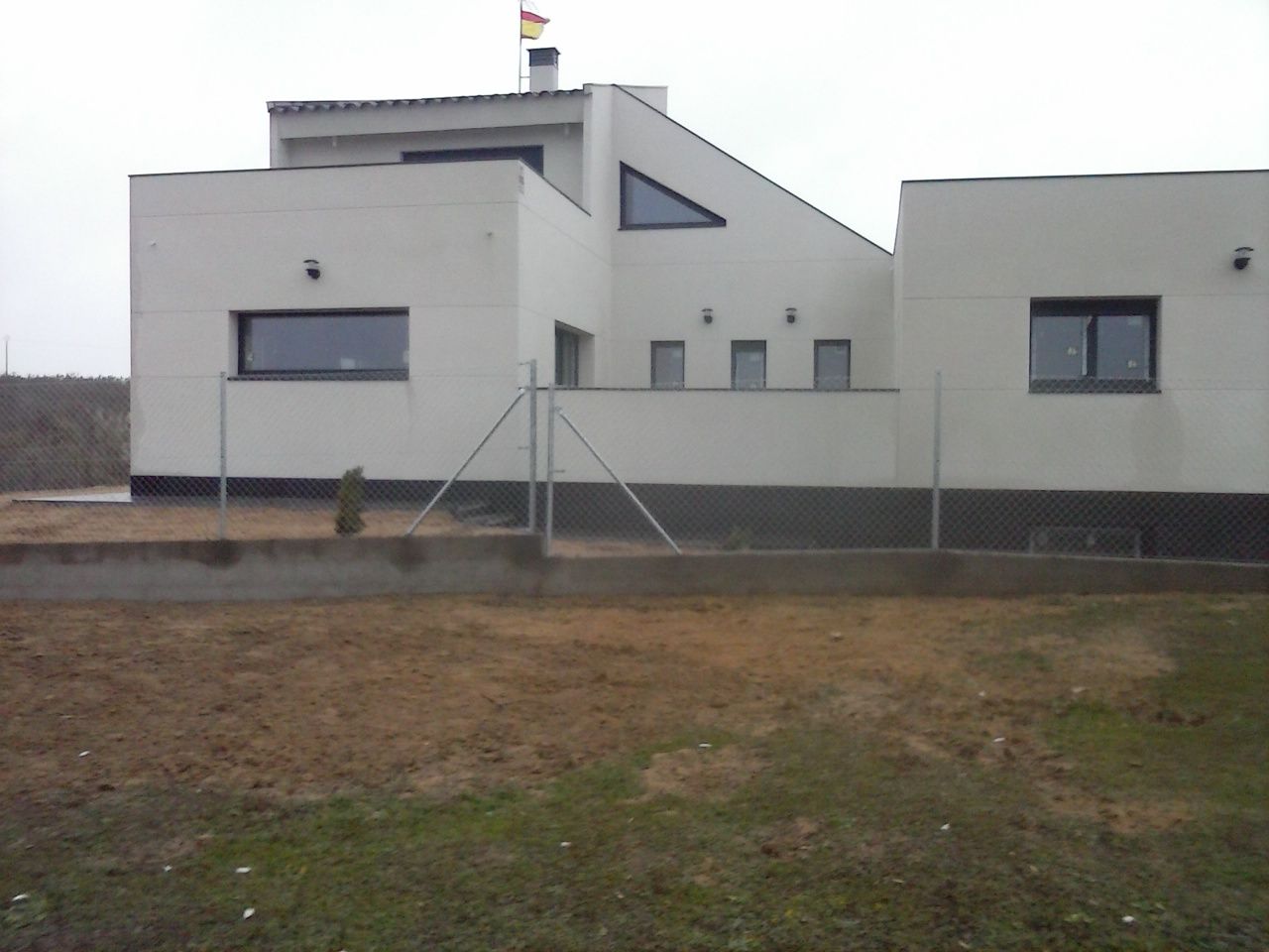 Casa blanca moderna con una bandera española en el tejado, detrás de una valla de alambre.