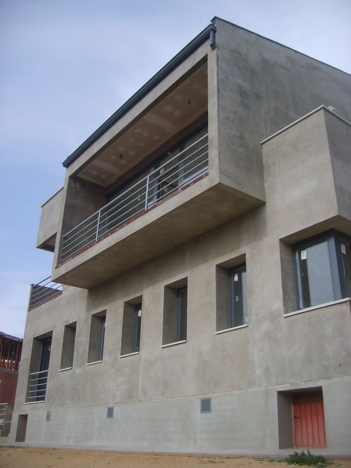 Edificio moderno gris con balcón rectangular y ventanas, bajo un cielo azul.